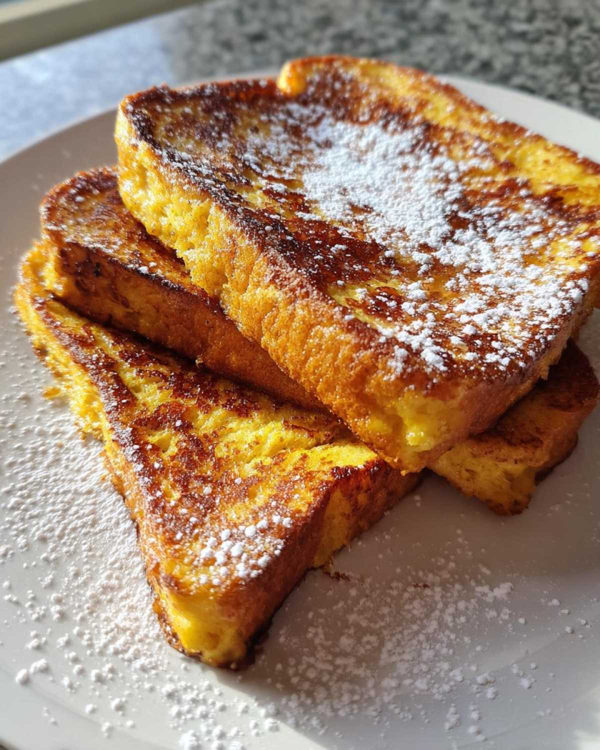 Close-up of three golden brown slices of easy french toast stacked and dusted heavily with powdered sugar.