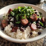 A white bowl filled with white rice, black beans and rice, sliced sausage, and topped with fresh cilantro.