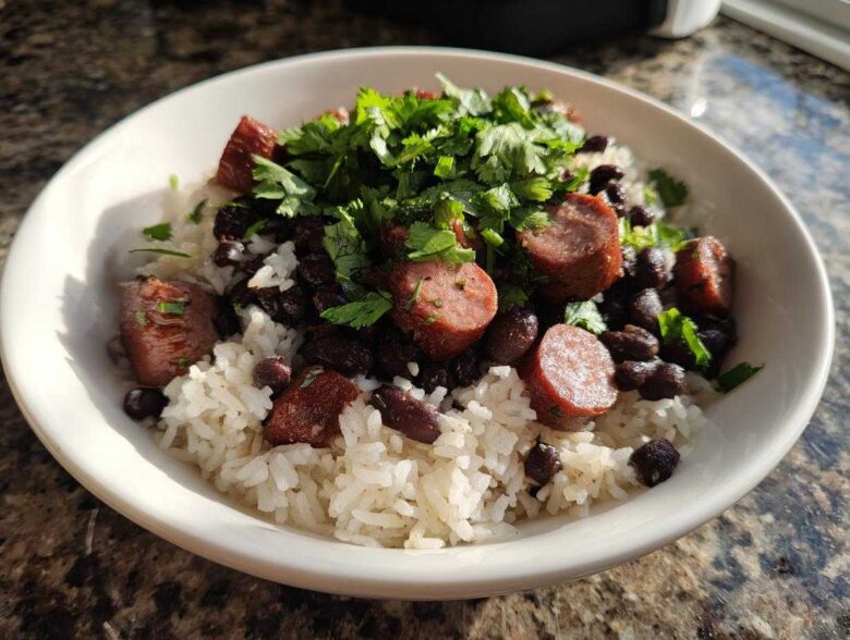 A white bowl filled with white rice, black beans and rice, sliced sausage, and topped with fresh cilantro.