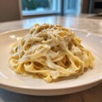 A serving of fettuccine pasta generously coated in creamy homemade alfredo sauce and topped with black pepper.