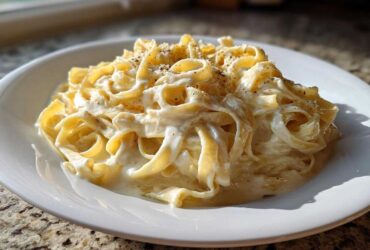 A close-up of fettuccine pasta generously coated in creamy homemade alfredo sauce and topped with black pepper.