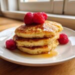 A stack of three fluffy almond flour pancakes drizzled with syrup and topped with fresh raspberries.