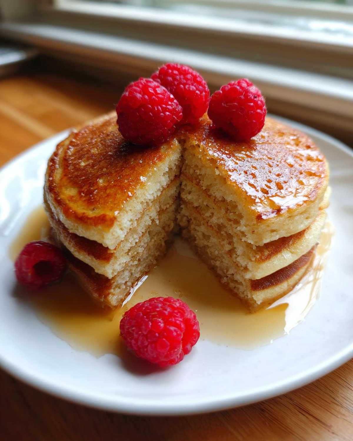 A fluffy stack of almond flour pancakes, cut in half, drizzled with syrup and topped with fresh raspberries.