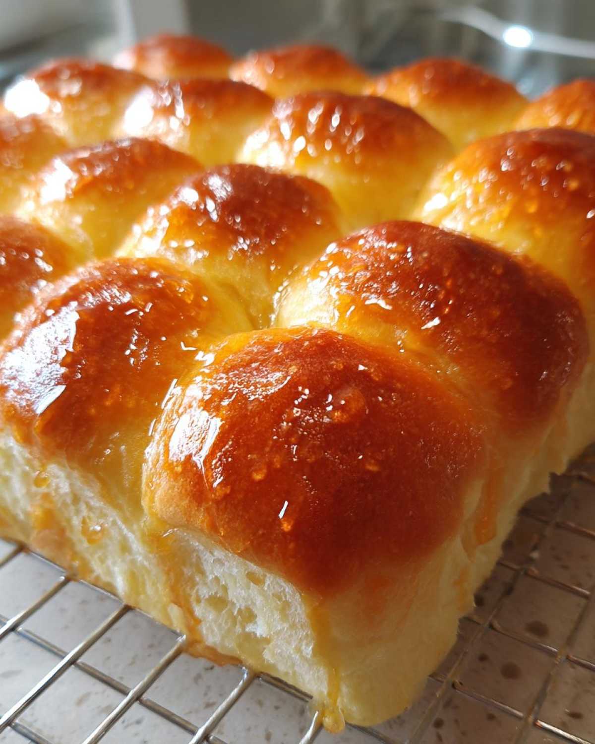 A close-up view of freshly baked, golden brown dinner rolls covered in a shiny, buttery glaze, resting on a cooling rack.