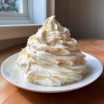 A large, beautifully piped mound of fluffy white cream cheese frosting sitting on a small white plate.