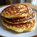 A close-up stack of three golden-brown, fluffy greek yogurt pancakes on a white plate.