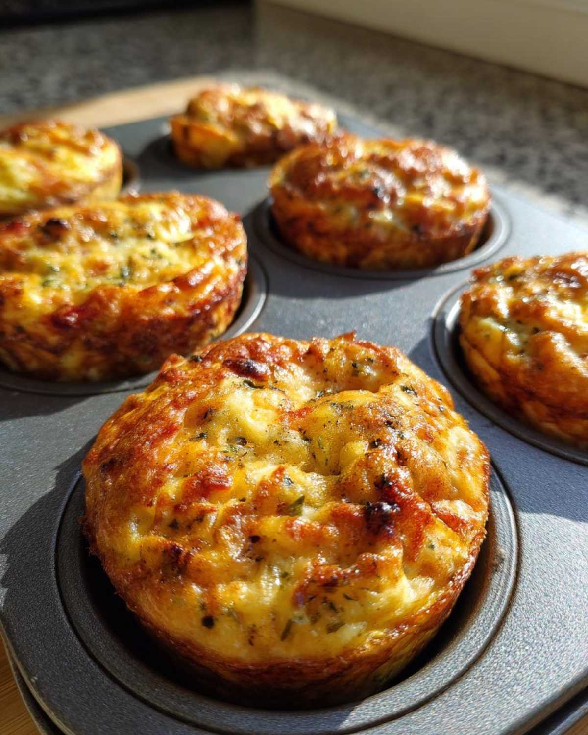 Close-up of golden brown Egg Muffin Cups baked in a dark metal muffin tin, glistening in the sunlight.