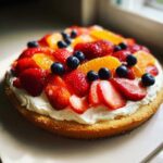 A round fruit pizza sugar cookie base topped with whipped cream, strawberries, blueberries, and orange slices.
