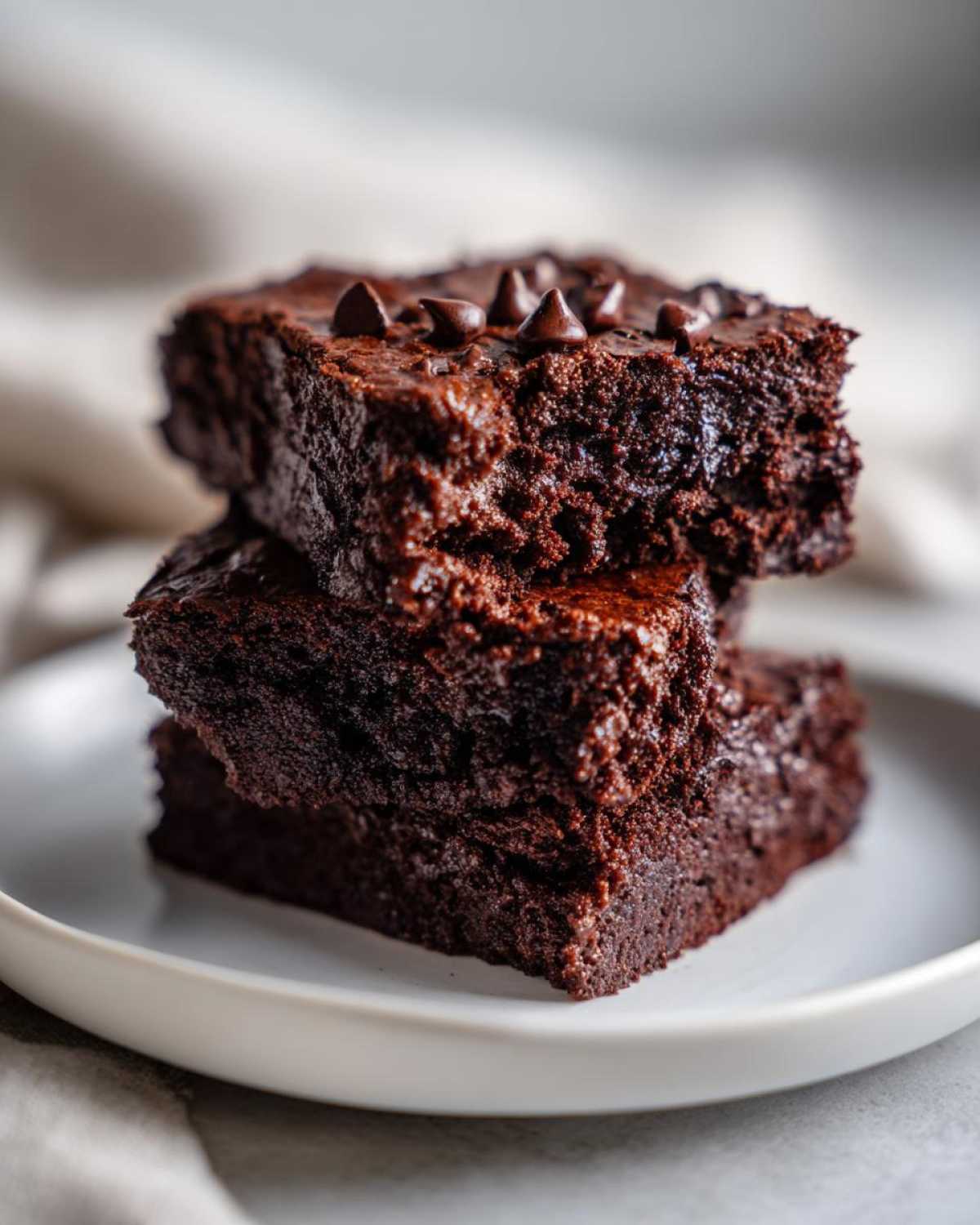 Close-up of three stacked, fudgy sweet potato brownies topped with chocolate chips on a white plate.