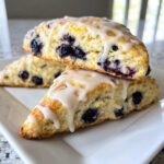 Close-up of three delicious blueberry scones stacked on a white plate, topped with a thick lemon glaze.