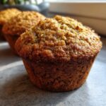 A close-up of a perfectly baked, golden-brown honey bran muffin sitting on a gray surface, with more muffins blurred in the background.