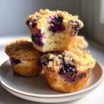 Close-up of moist blueberry muffins stacked, showing the bright purple interior and crunchy streusel topping.
