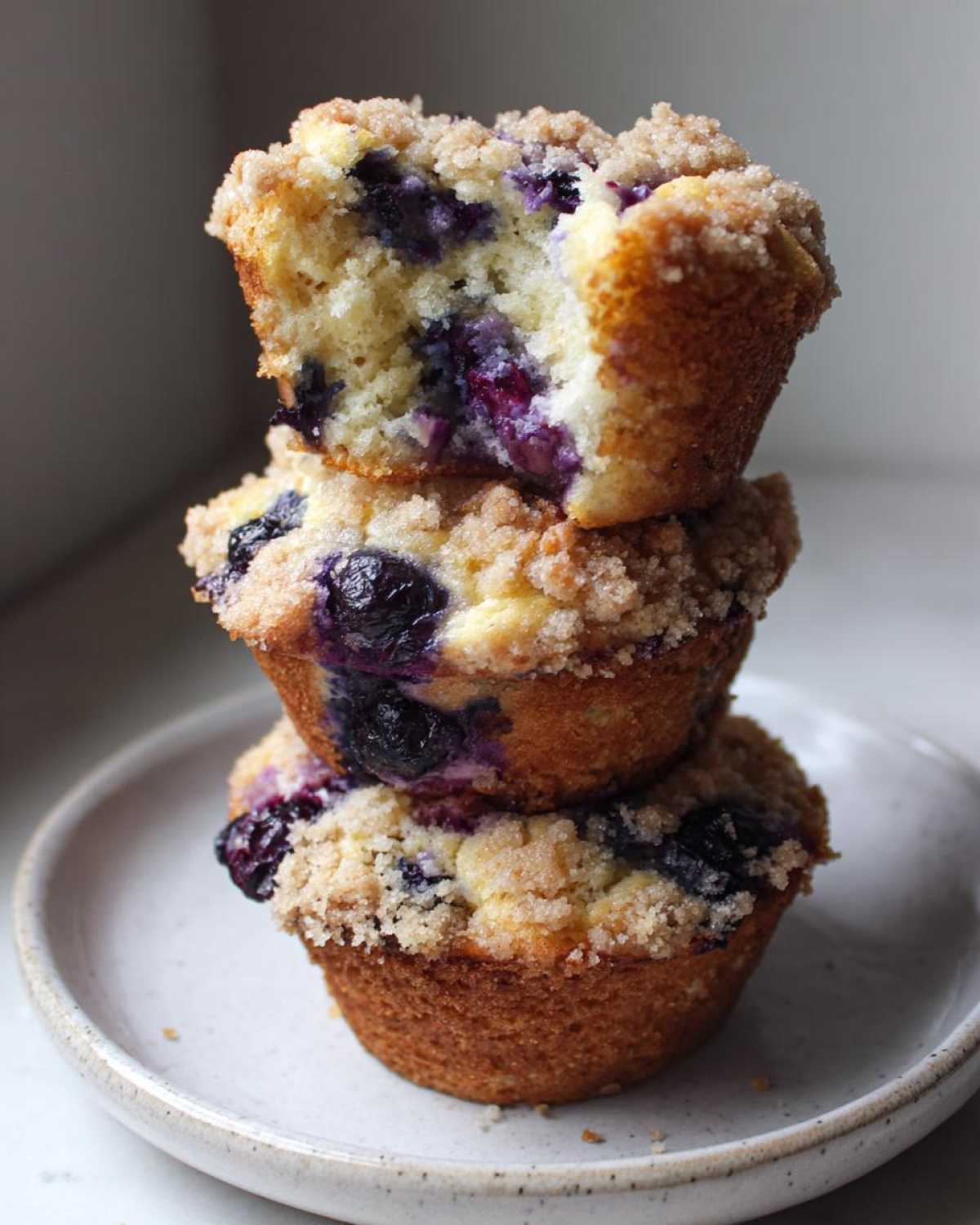 A stack of three moist blueberry muffins, topped with streusel, with the top one broken open to show the interior.
