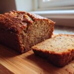 A close-up of a moist sugar free banana bread loaf, partially sliced on a wooden cutting board.