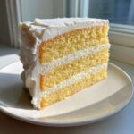Close-up of a moist, three-layer slice from the best vanilla cake recipe with thick white frosting.