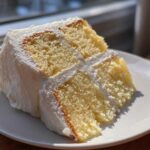 A close-up of a perfectly moist slice from the best vanilla cake recipe, layered with white frosting.