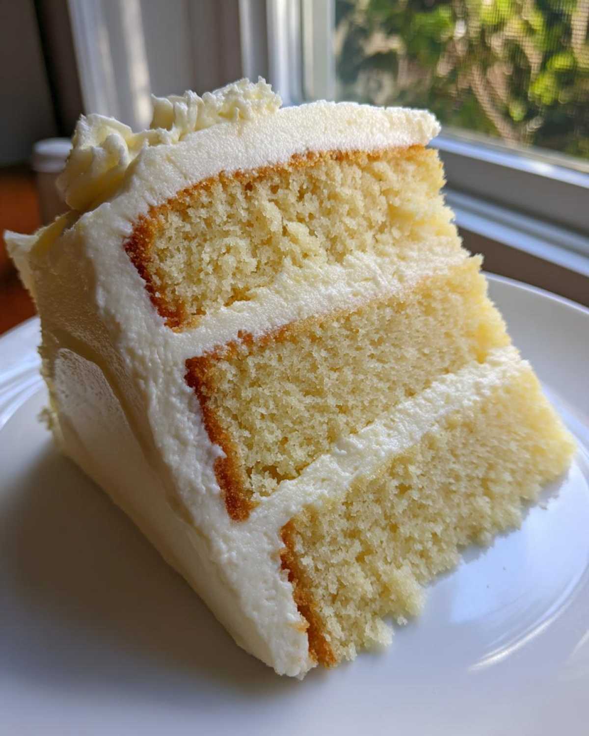 Close-up of a moist slice from the best vanilla cake recipe, showing three layers and thick white buttercream frosting.