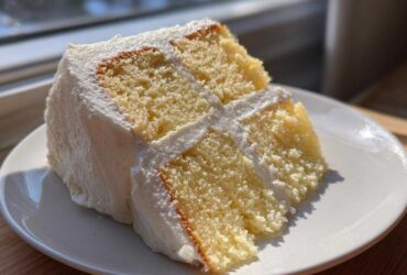 A close-up of a perfectly moist slice from the best vanilla cake recipe, layered with white frosting.