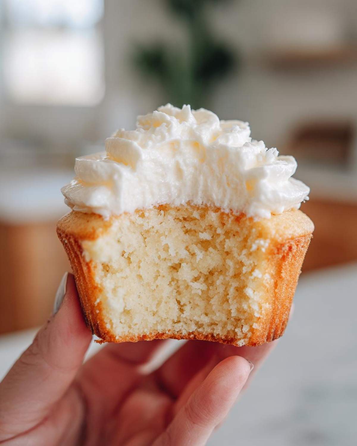 Close-up of a hand holding a vanilla cupcake cut in half, showing the soft, moist crumb and thick white frosting.