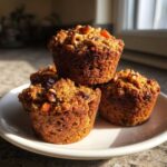 A stack of four golden brown morning glory muffins topped with nuts and dried fruit, sitting on a white plate.