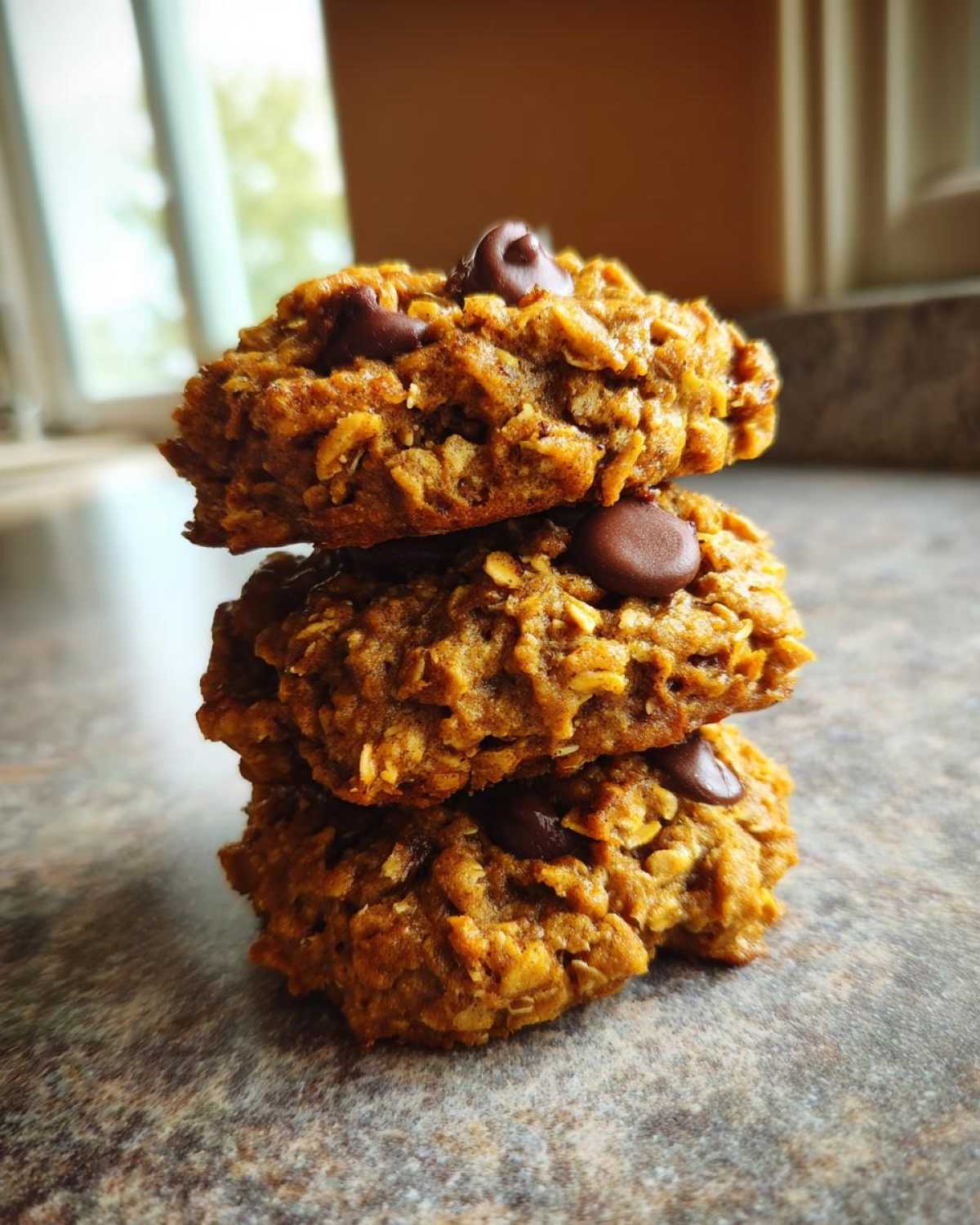 Three oatmeal chocolate chip breakfast cookie stacked vertically on a speckled countertop.