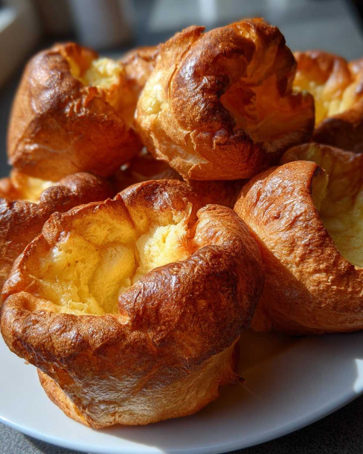 Close-up of several golden-brown, puffy yorkshire pudding pastries piled on a white plate.