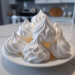 A pile of glossy, perfectly piped white meringue cookies stacked on a small white plate.