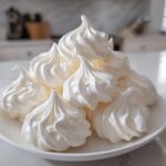 A pile of perfectly piped, glossy white meringue cookies stacked on a small white plate.
