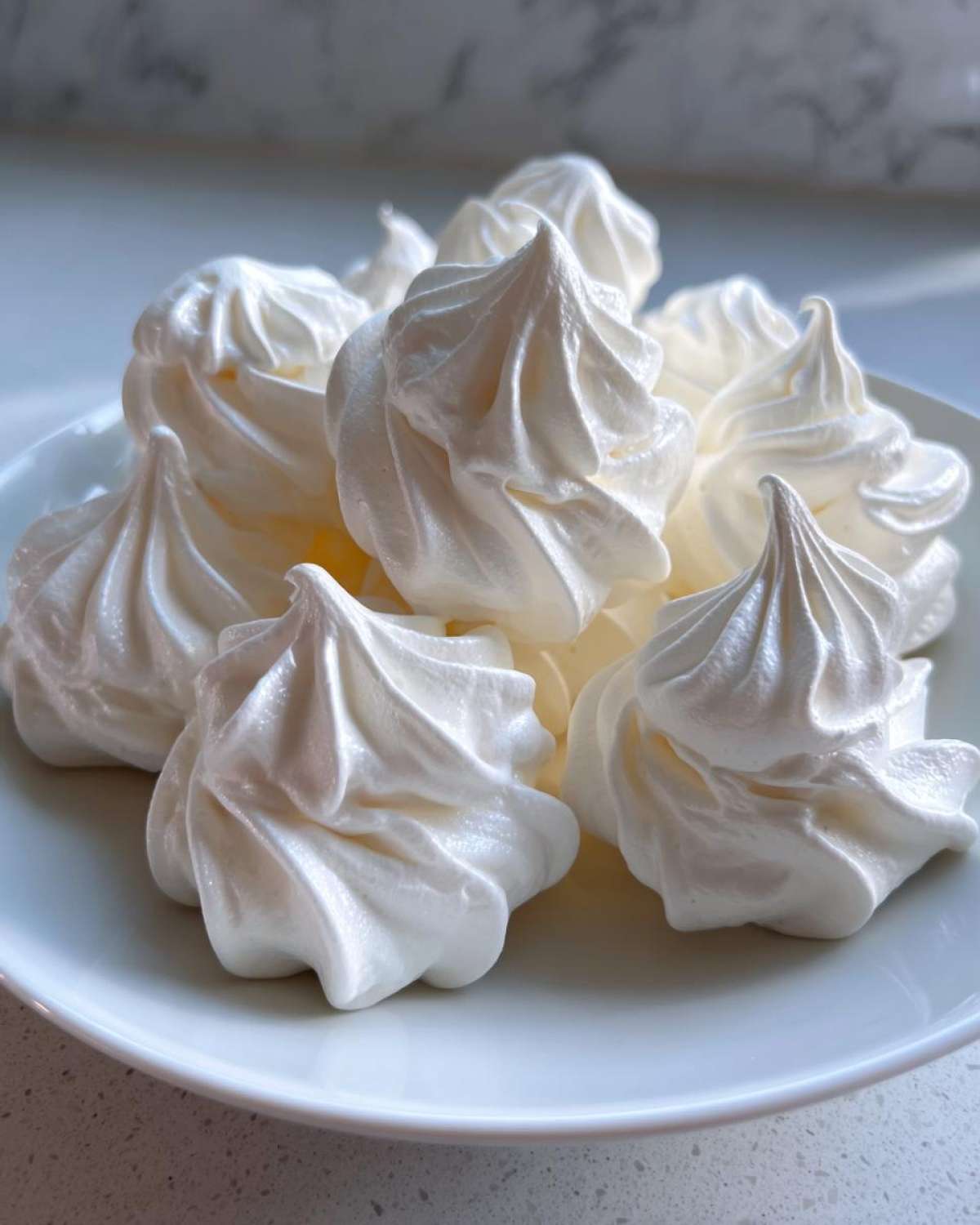 A close-up of several perfectly piped, glossy white meringue cookies piled on a simple white plate.