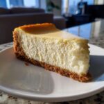 A close-up, sunlit slice of creamy Protein Cheesecake with a thick graham cracker crust on a white plate.
