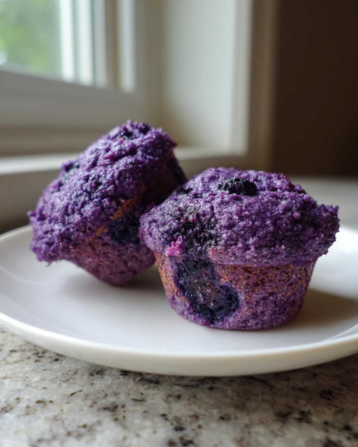 Two vibrant purple blueberry protein muffins sitting on a white plate near a window.