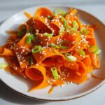 Close-up of a vibrant raw carrot salad made with shaved carrot ribbons, topped with white sesame seeds and chopped green onions.