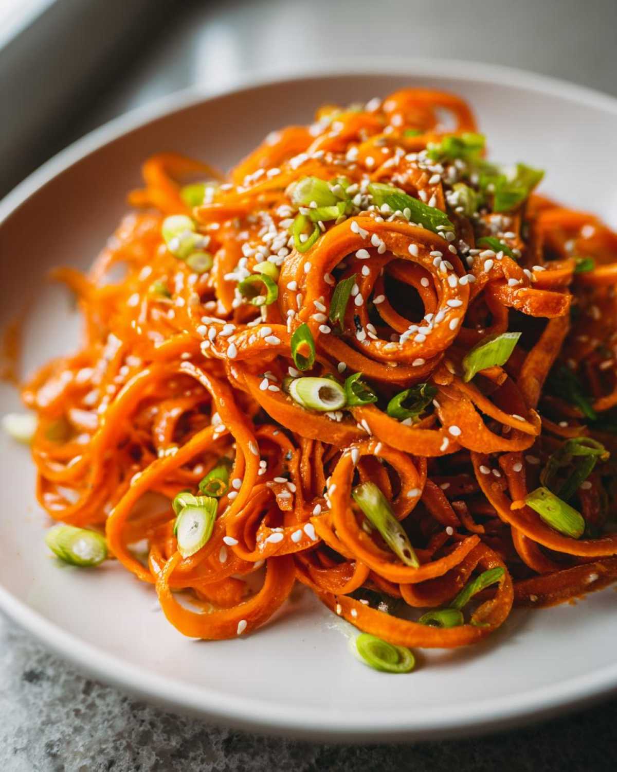 Close-up of vibrant orange spiralized raw carrot salad topped with white sesame seeds and sliced green onions.