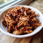 A close-up shot of tender, shredded crock pot pulled pork generously coated in barbecue sauce in a white bowl.