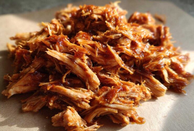 A close-up, appetizing pile of shredded, saucy slow cooker pulled pork resting on brown paper.