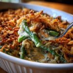 A spoonful of creamy green bean casserole topped generously with crispy fried onions being lifted from a white baking dish.
