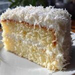 Close-up of a moist slice of coconut cake layered with white frosting and topped with shredded coconut.