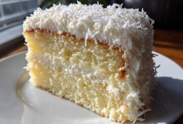 Close-up of a moist slice of coconut cake layered with white frosting and topped with shredded coconut.
