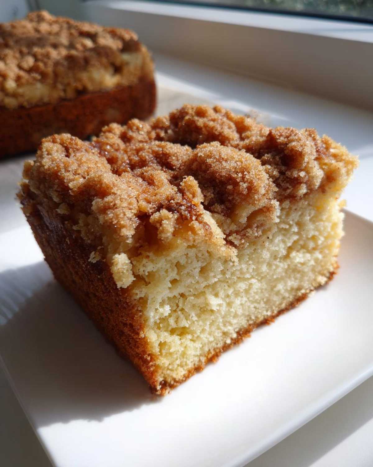 Close-up of a moist slice of coffee cake with a thick, sugary cinnamon streusel topping on a white plate.