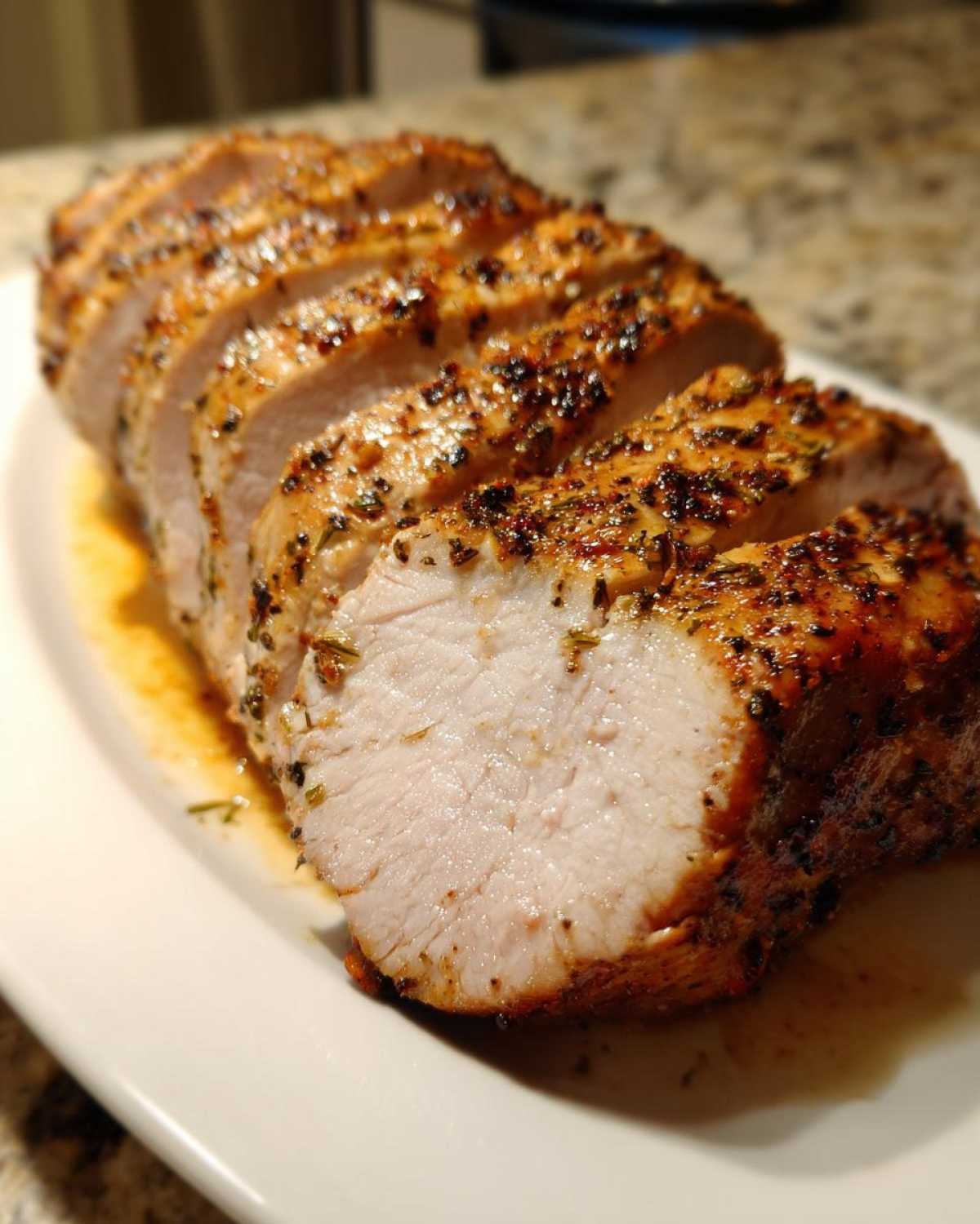 Close-up of a juicy, sliced pork loin roast with a dark herb crust resting in its juices on a white platter.