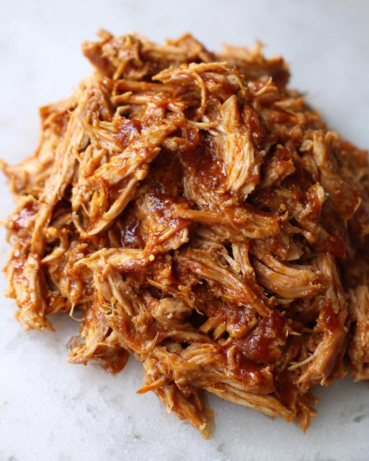 A close-up pile of shredded, saucy Slow cooker pulled pork resting on a white marble surface.