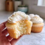 Close-up of a hand holding a soft vanilla cupcake with white frosting, showing the fluffy interior after a bite was taken.
