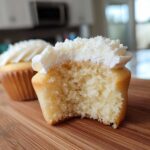 A vanilla cupcake cut in half showing its soft, moist crumb and white vanilla frosting. Another vanilla cupcake is visible in the background.