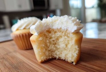 A vanilla cupcake cut in half showing its soft, moist crumb and white vanilla frosting. Another vanilla cupcake is visible in the background.