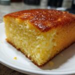 Close-up of a moist, golden slice of sourdough cornbread with a shiny, browned top, resting on a white plate.