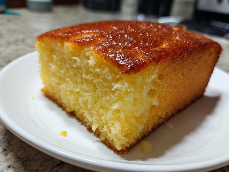 Close-up of a moist, golden slice of sourdough cornbread with a shiny, browned top, resting on a white plate.