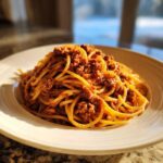 A close-up of spaghetti generously topped with rich bolognese sauce, served on a white plate.