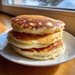 A stack of three thick, fluffy pancakes with beautifully browned tops served on a white plate.