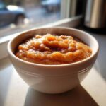 Close-up of a white bowl filled with warm, spiced applesauce steaming slightly by a window.