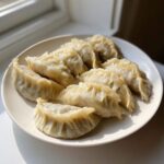 A plate filled with freshly steamed Traditional Chinese Dumplings, showing crimped edges, set near a window.